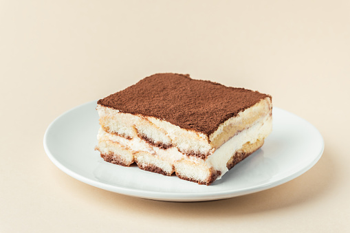 Homemade tiramisu cake on white plate against beige background. Traditional Italian no-bake dessert made of savoiardi, filled with mascarpone cheese, coffee espresso and sprinkled with cocoa powder