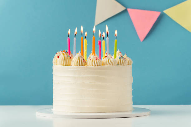 istock Birthday cake with white cream cheese frosting decorated with multicolored lit candles on a blue background. Happy Birthday concept. Tradition of making a wish while blowing out candles on a cake 1454670841