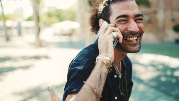 istock Attractive italian guy with ponytail and stubble is using mobile phone sitting on bench at old buildings background. Stylish man emotionally talking on smartphone 1454675110