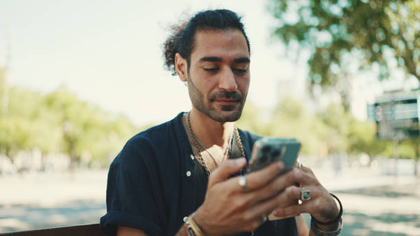 istock Attractive italian guy with ponytail and stubble is using mobile phone sitting on bench at old buildings background. Stylish man writes message, views photo on smartphone 1454675175