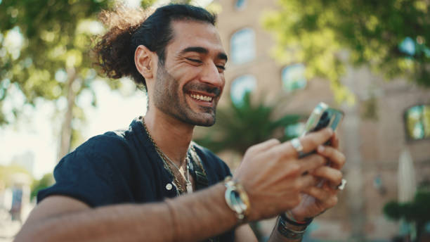 istock Attractive italian guy with ponytail and stubble is using mobile phone sitting on bench at old buildings background. Stylish man writes message, views photo on smartphone 1454675184