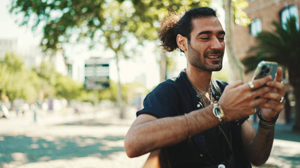 istock Attractive italian guy with ponytail and stubble is using mobile phone sitting on bench at old buildings background. Stylish man writes message, views photo on smartphone 1454675204
