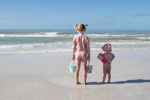 Two sisters, in pink UV clothes, standing on a white beach, watching the waves of the ocean. A blue sky and horizon in the background. The older girl wearing buckets in each hand, the younger girl wearing a flotation device. A hot sunny day. Copy space.