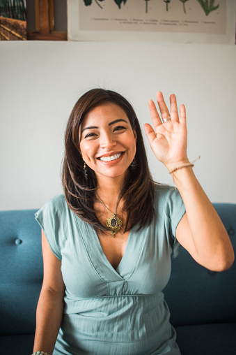 vertical portrait of a woman waving and smiling at the camera sitting in her living room at home