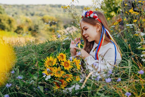 Little girl wearing Ukrainian national dress and wreath of flowers outdoor. Portrait of teenager in authentic folk dress with embroidery on the meadow.