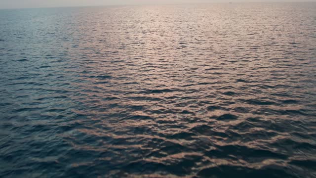 The sun beyond the horizon. Pink clouds and rose lights of the sunrise reflected in the surface of the water. Sunrise at sea. Sunset at sea. Backwards dolly shot with tilt down camera movement. At the end a close-up of the water surface. Version 7
