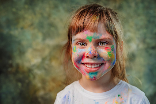 Little girl's face covered with holi colors. Portrait of happy smiling toddler with colotful paintings on the skin.