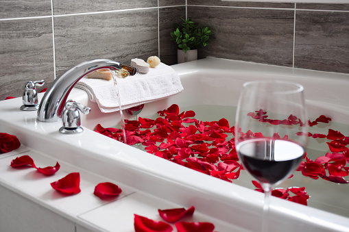 Glass of red wine on the edge of bathtub filled with red rose petals