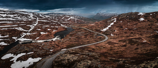 Lonesome road through the landscape of Hardangervidda National Park in Norway, snow fields and dark clouds in the sky, from above