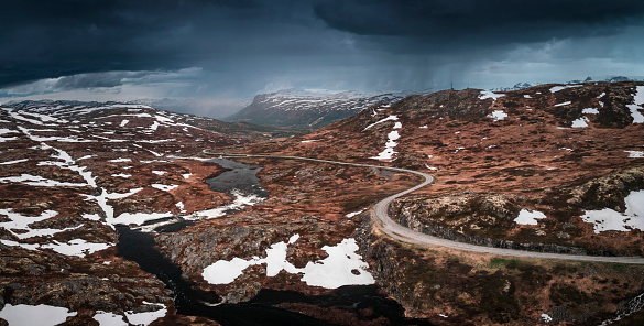 Lonesome road through the landscape of Hardangervidda National Park in Norway, snow fields and dark clouds in the sky, from above