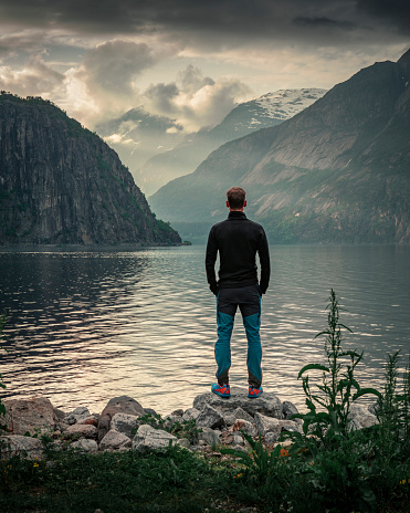 Man standing at waterfront of lake in the mountain landscape Eidfjord in Norway, looking into the fjord, clouds in the sky during sunset
