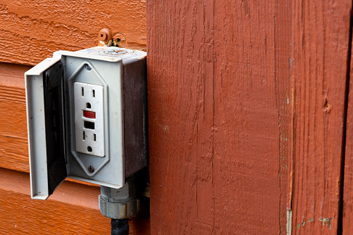 An image of an old outdoor electrical outlet on the exterior wall of a reddish brown building.