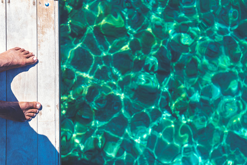 Man standing on a jetty showing his feet and the water. The water is clear and transparent and turquoise blue.