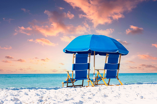 A set of blue beach chairs sitting on the sand facing the ocean horizon with a dramatic sunset.