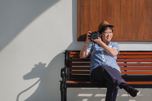 Portrait of happy Asian adult female tourist in vintage style holding digital camera and looking at camera while sitting on wooden bench outside of building
