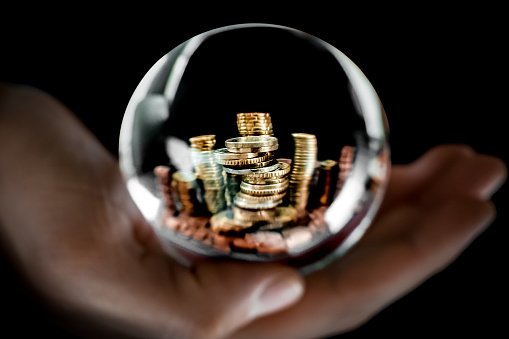Imagen simbólica de una bola de cristal redonda en una mano humana con ahorros de dinero en efectivo en forma de varias pilas de monedas de euro metálicas brillantes dentro del momento de la inflación con espacio de copia en la parte superior. photo