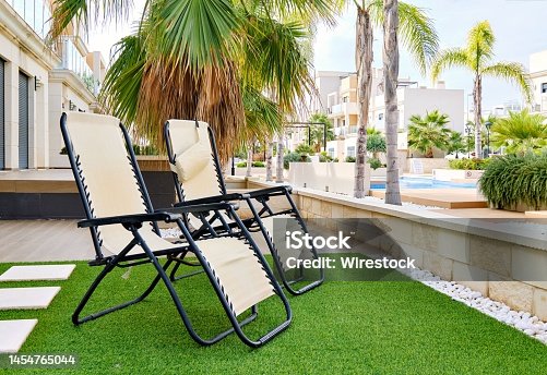 istock Two lounge chairs on artificial grass with modern apartments and a swimming pool in the background 1454765044