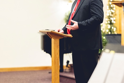 A male wearing a black suit and preaching holding onto a pulpit