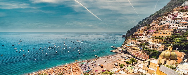 The Amalfi coast with beach, hillside architecture, jets leave white trail in cloudy sky, Positano, Italy