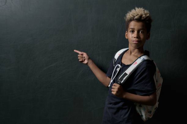 istock Black schoolboy wearing a backpack pointed his finger at dark grey background with copy space 1454768027