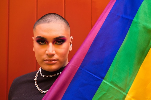 A Hispanic boy with vibrant makeup holding gay pride flag against red wooden door