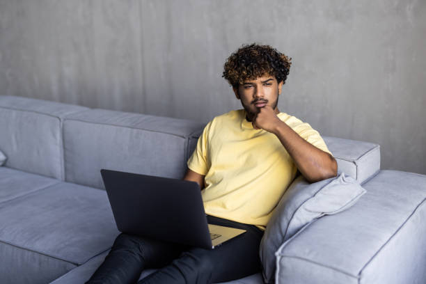 istock Smiling ethnic indian man sitting at home on sofa using laptop looking at computer working or elearning online, watching movies or having virtual chat remote videocall. 1454777231