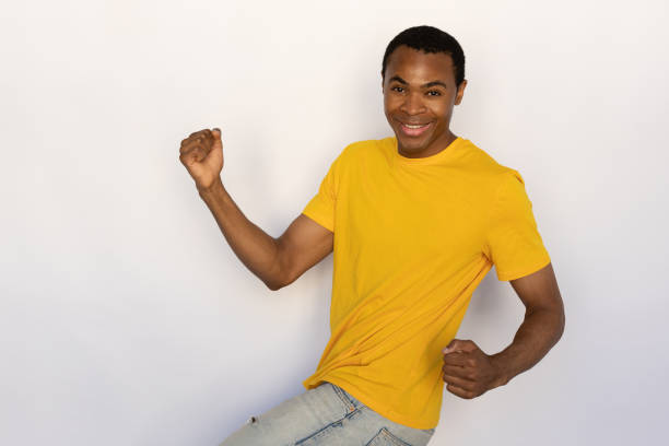 Overjoyed young man celebrating victory. Male African American model with brown eyes and short black haircut in yellow T-shirt showing winning gesture with raised fists. Achievement, success concept