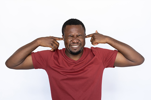 Irritated young man covering his ears with fingers. Male African American model with closed eyes, short black haircut and beard in casual clothes protecting his ears from loud sound. Silence concept