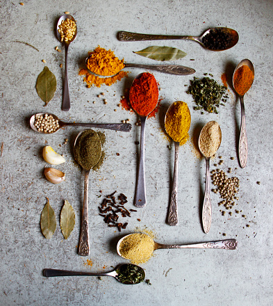 A top view of various spices in metal spoons with gray marble background.
