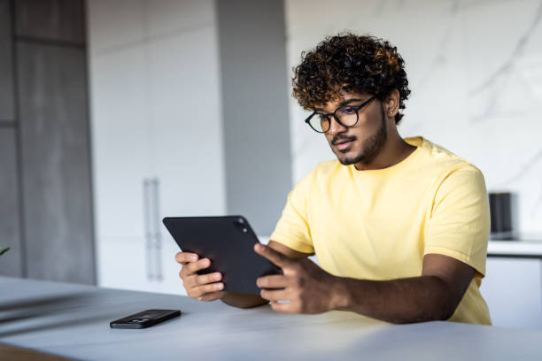 istock Young handsome indian man drinking coffee and looking at digital tablet in the kitchen. 1454779809