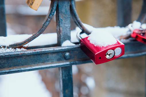 Red padlock is hanging on the metallic bars fence covered with snow