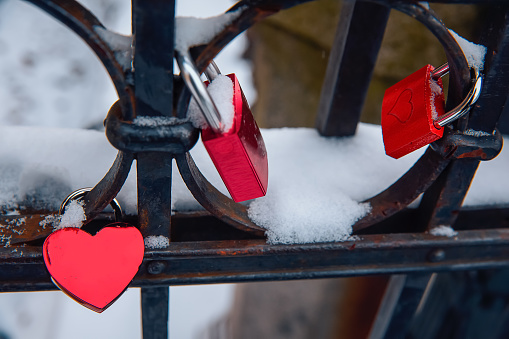 Red heart love shaped padlocs are hanging on the metallic bars fence covered with snow