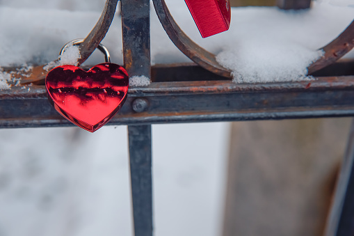 Red heart love shaped padlocs are hanging on the metallic bars fence covered with snow