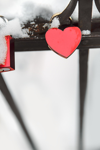 Red heart love shaped padlocs are hanging on the metallic bars fence covered with snow