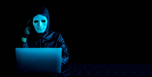 istock Hacker in mask and hoodie using a laptop to browse the net and serious face thinking about question 1454781643