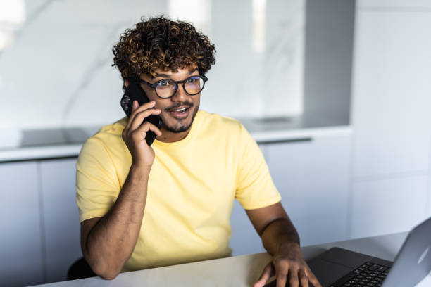 istock Happy indian guy having conversation on phone, using laptop at kitchen 1454782952