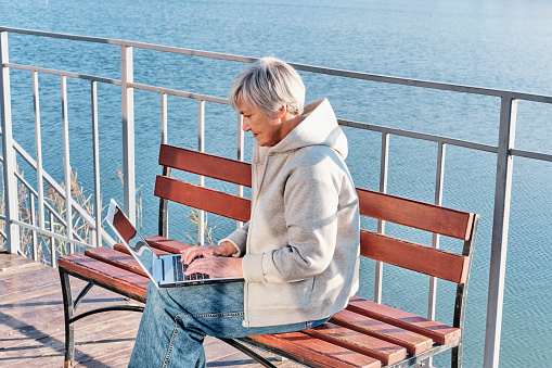 Senior woman with short gray hair, wear jeans and hoodie, sitting on bench near lake, using laptop. Working online, using some online services, online shopping. Technology, elderly, concept