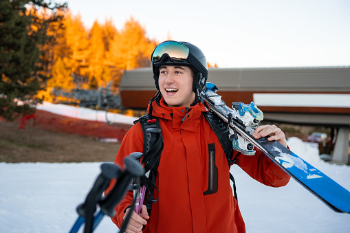 Young boy wearing ski clothes and carrying ski equipment on his shoulders for skiing. He is walking, talking, smiling. Finishing the ski day coming home