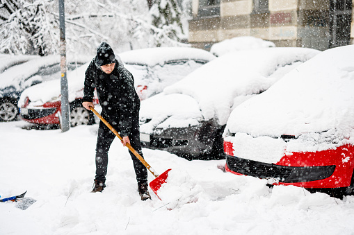 A Man with Snow Shovel is Shoveling a Snow in Front of Car During a Cold Snowy Winter Day.