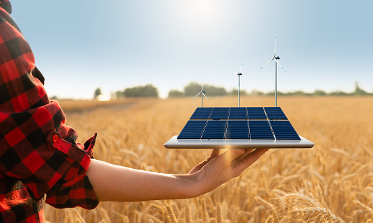 Farmer is holding a digital tablet with solar panels and wind turbines. Sustainable farming concept