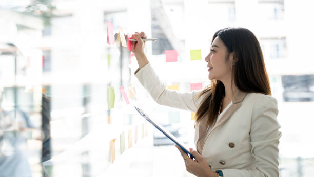 istock Young smiley attractive, businesswoman using post it notes in glass wall to writing strategy business plan to development grow to success 1454805521