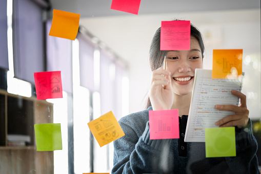 Young smiley attractive, businesswoman using post it notes in glass wall to writing strategy business plan to development grow to success
