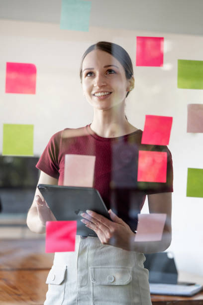 istock Young smiling caucasian businesswoman mentor leader write tasks creative ideas on sticky post it notes on glass wall. 1454806557