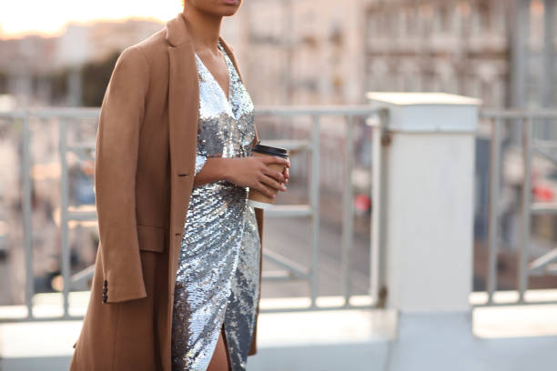 Woman in shiny silver sequin dress and beige coat with take away coffee cup standing on roof terrace
