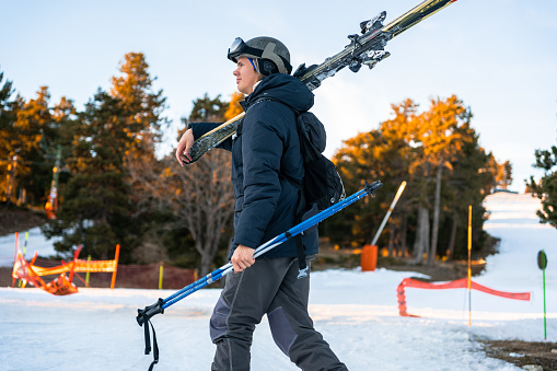 Young boy wearing ski clothes and carrying ski equipment on his shoulders for skiing. He is looking away and carruying the skii sticks on the left hand. Snow can be seen. Finishing the ski day coming home