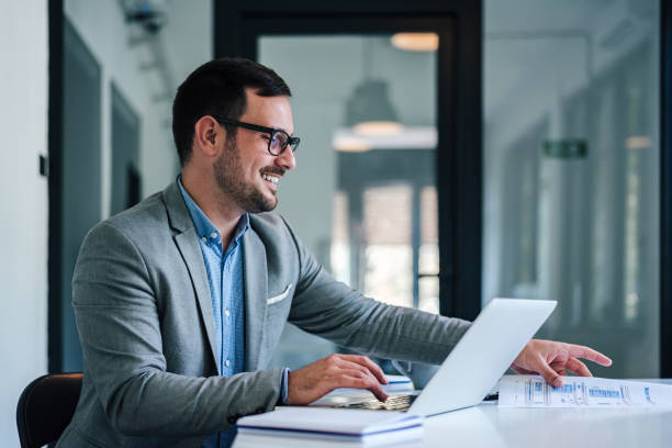 istock Relaxed caucasian banker, taking papers from his desk, using laptop. 1454814596