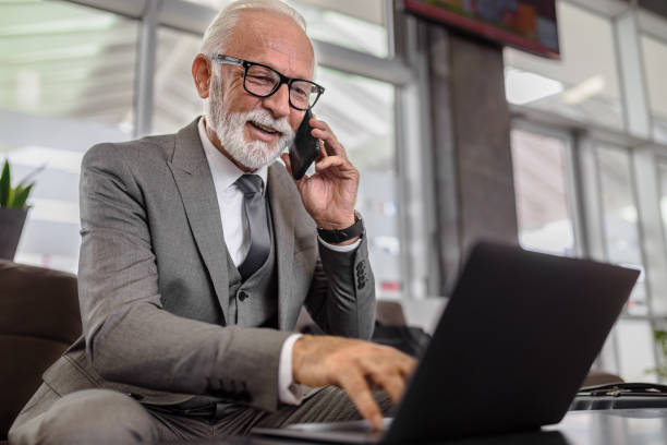 istock Smiling elderly male professional in suit using laptop while talking on smart phone at the office 1454814934