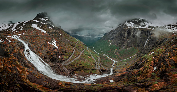 Mountain road Trollstigen winding through landscape with waterfall and valley of Trollveggen in Norway, dramatic sky with dark clouds, from above