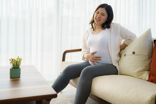 Asian pregnant woman suffering from backache, sitting on couch, holding belly, touching back. Expectant mother tired of overweight, having health problems, feeling lumbago muscles pain.