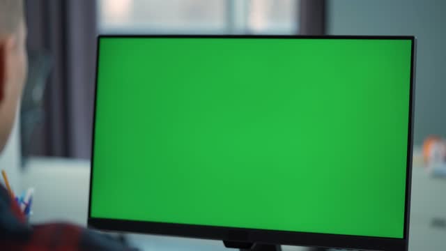 Young Man Working At Computer With Green  Mock Up Screen  Sitting At Desk in Office